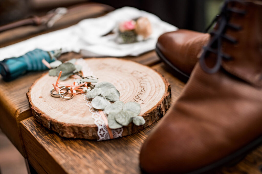 Zwei Eheringe liegen auf einer Holzscheibe, die mit Spitze und Eukalyptusblättern dekoriert ist. Im Hintergrund sind ein Paar braune Lederschuhe und weitere Hochzeitsaccessoires zu sehen.