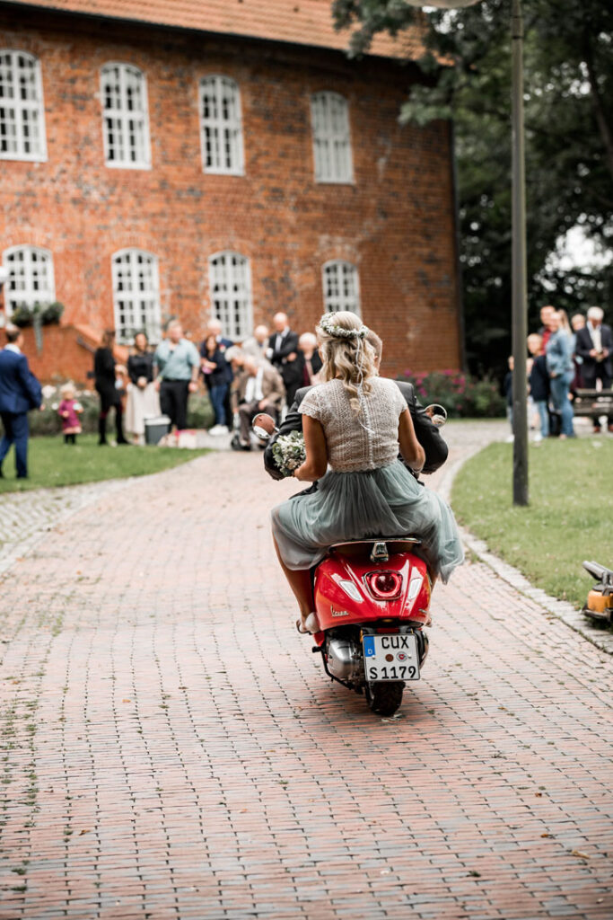 Eine Braut in einem weißen Spitzenkleid fährt auf einem roten Roller, während Gäste im Hintergrund stehen.