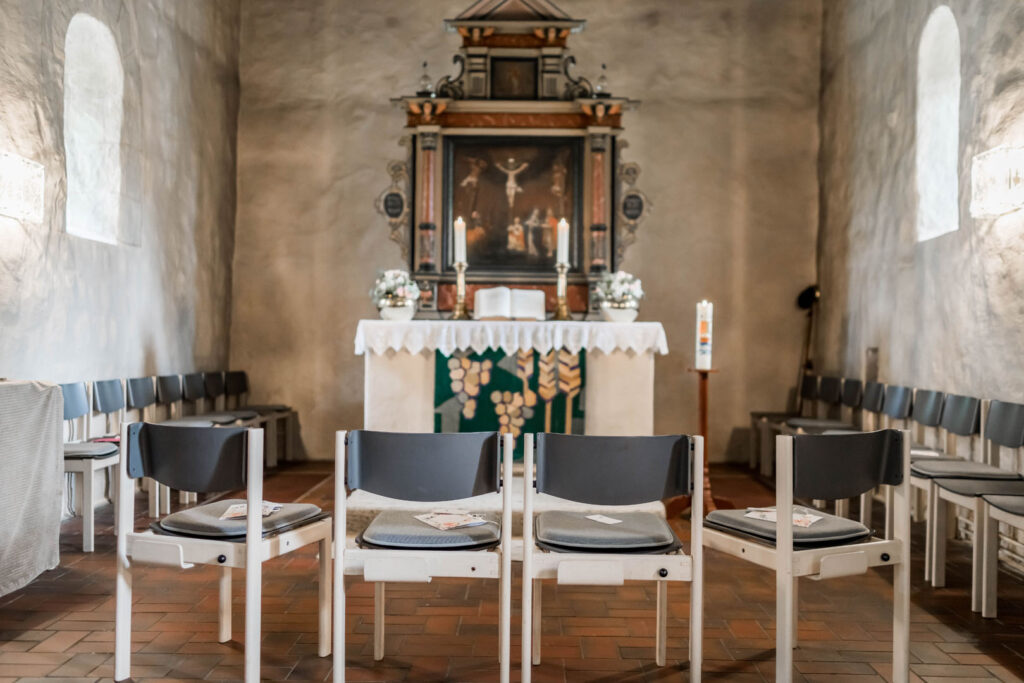 Blick auf den Altar einer Kirche mit drei Stühlen im Vordergrund und einem Gemälde im Hintergrund.