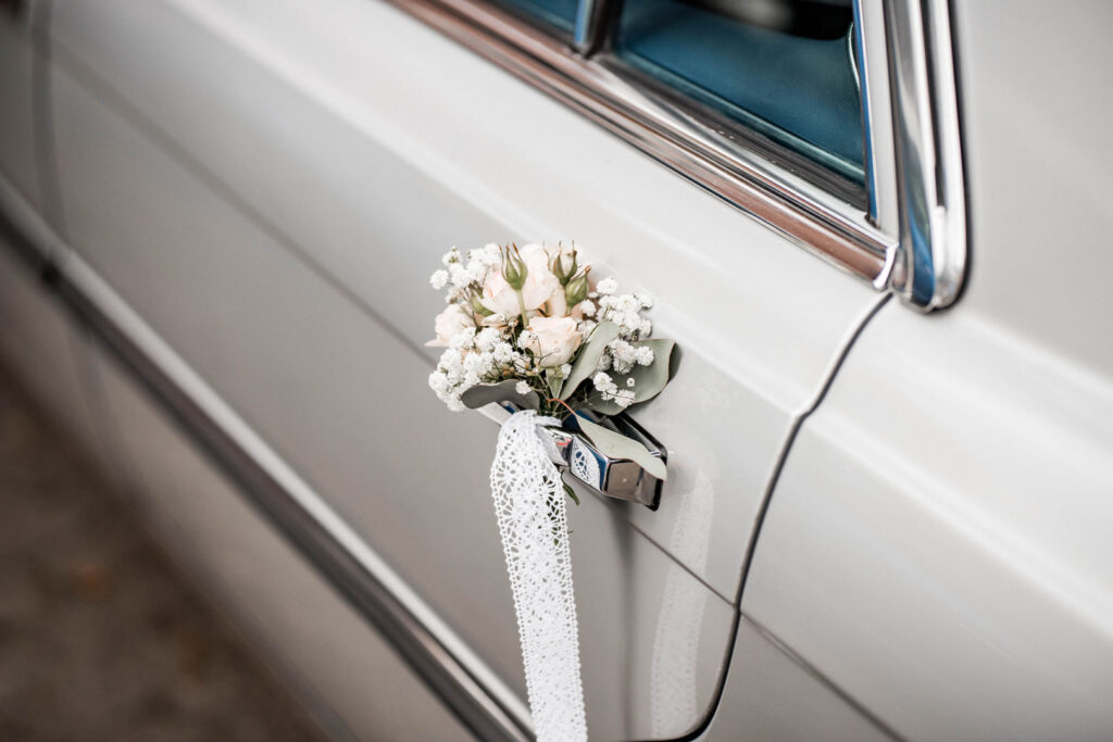 Ein kleiner Blumenstrauß mit weißen und rosa Blüten ist an einem Türgriff eines silbernen Autos befestigt.