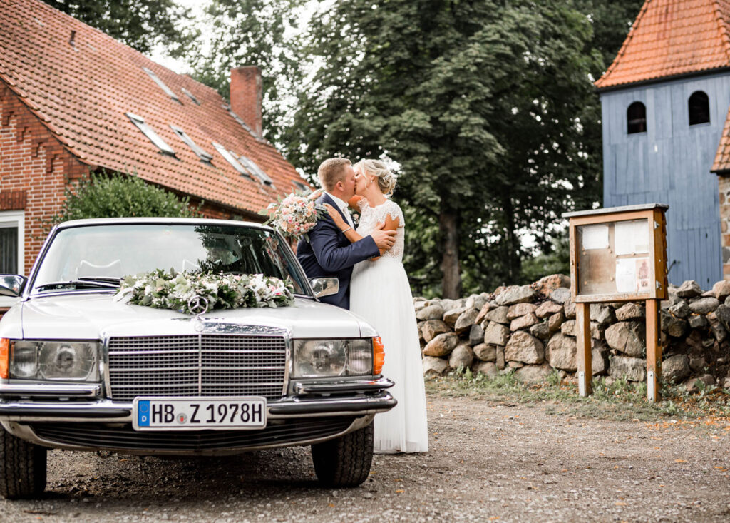 Ein Brautpaar küsst sich vor einem silbernen Oldtimer, der mit Blumen geschmückt ist. Im Hintergrund sind ein Backsteingebäude und eine Kirche zu sehen.