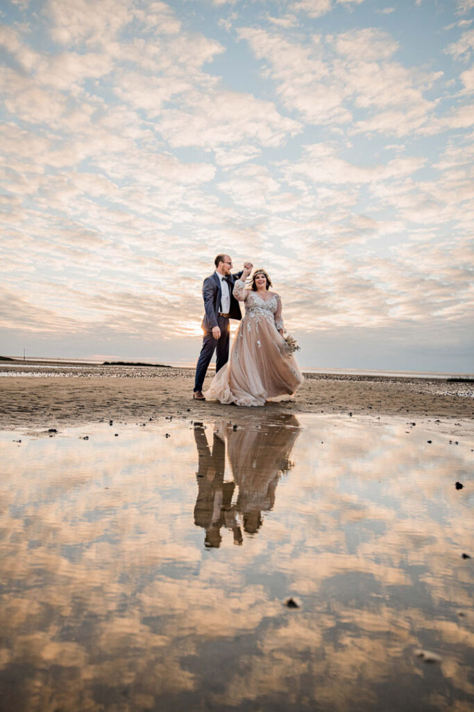 Kira und Sönke posieren am Strand. In der Pfütze vor ihnen reflektiert sich der wunderschöne Himmel.