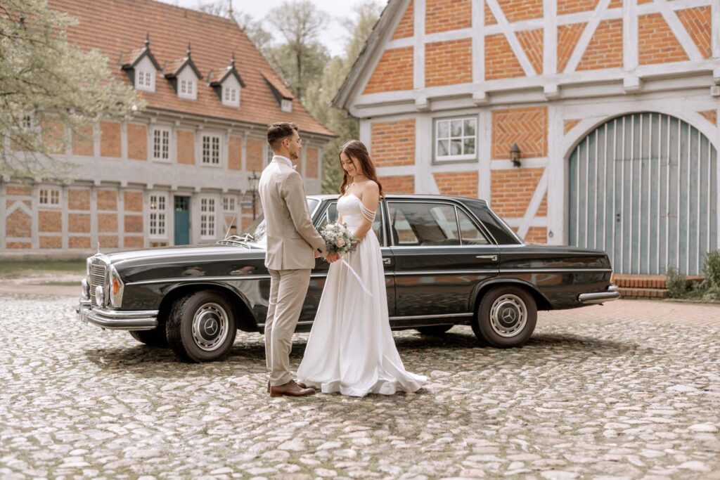 Christina und Kevin stehen am Tag ihrer Hochzeit vor einem Mercedes auf dem Hof eines idyllischen Landhauses.