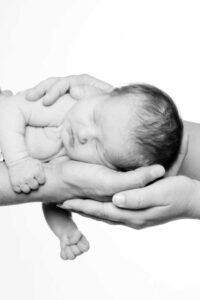 Ein neugeborenes Baby wird behutsam von den Händen der Eltern gehalten, während es friedlich schläft. Das Bild ist in Schwarz-Weiß gehalten und hebt die zarten Details des Babys und die fürsorgliche Haltung der Hände hervor.