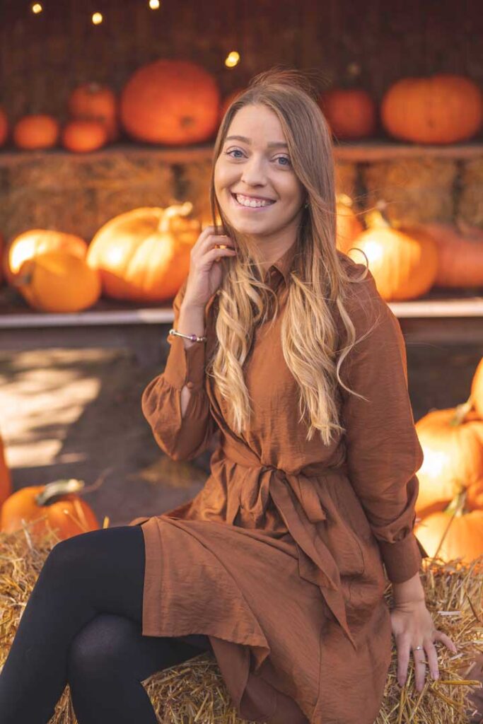 Eine junge Frau sitzt auf einem Heuballen, umgeben von Kürbissen, und lächelt in die Kamera. Sie trägt ein braunes Kleid, das perfekt zur herbstlichen Szene passt.