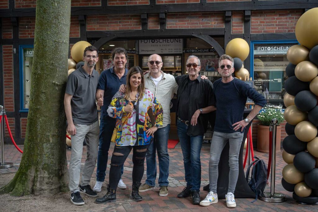 Eine Gruppe von sechs Personen steht vor einem Fotostudio mit einem Schild „Fotoscheune“. Sie posieren lächelnd und entspannt für die Kamera, umgeben von schwarzen und goldenen Luftballons.